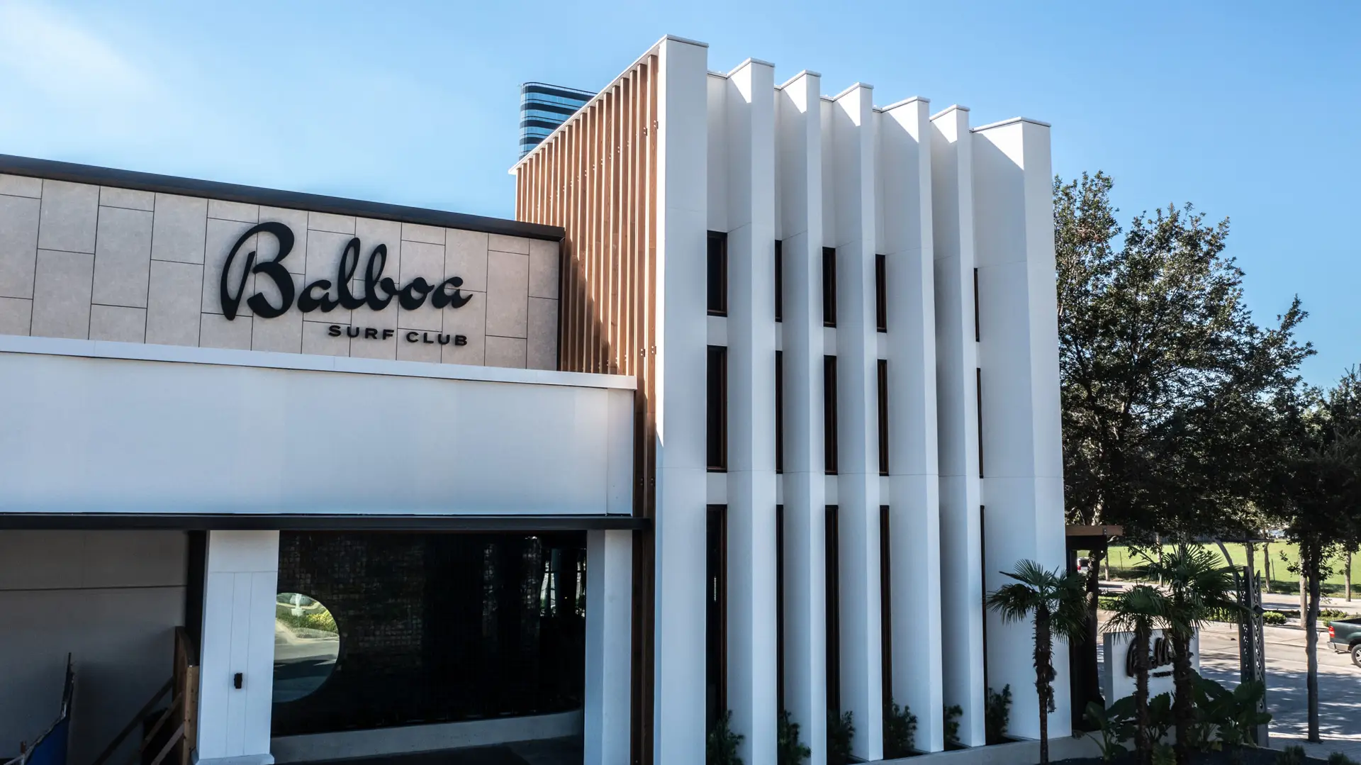 Balboa Surf Club exterior showing white vertical architectural panels, wood accents, black signage, and landscaped entry areas.