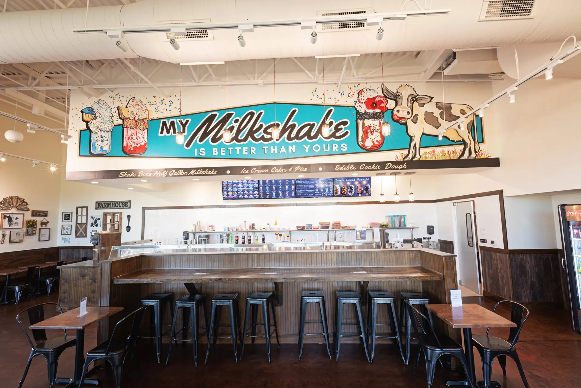 Interior view of The Yard Milkshake Bar showing the service counter, seating area, menu boards, and large milkshake-themed mural above the counter.